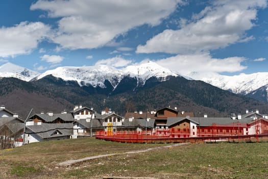 Snow-capped mountains tower over a quiet village with colorful houses and a playground in springtime - Rosa Khutor, Russia April 15, 2024.