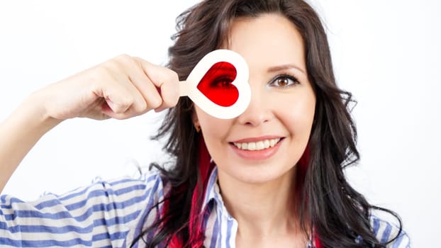 A young woman with long dark hair is smiling and holding a red heart-shaped lollipop to her right eye. She is wearing a blue and white striped shirt and has red earrings.