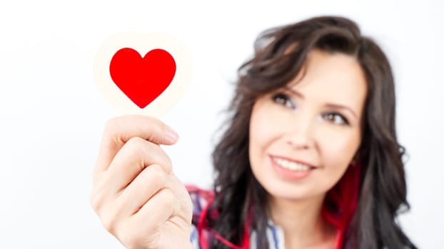 A young woman with long, dark hair smiles brightly as she holds a red heart-shaped paper cutout in her hand, wearing a blue and white striped shirt against a soft, out-of-focus white background.