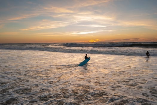 Two Surfer friends carving large breaking wave at colorful summer sunset. Reflective ocean waves carrying man on surfboard to tranquil beach on beautiful evening.