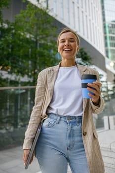 Smiling business woman drinking coffee during break time near office building and looks camera