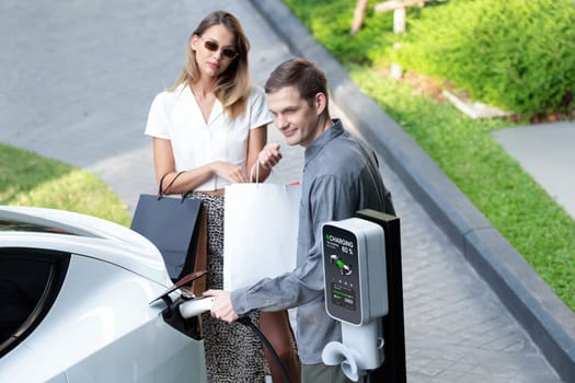 Young couple travel with EV electric car charging in green sustainable city outdoor garden in summer shows urban sustainability lifestyle by green clean rechargeable energy of electric vehicle innards