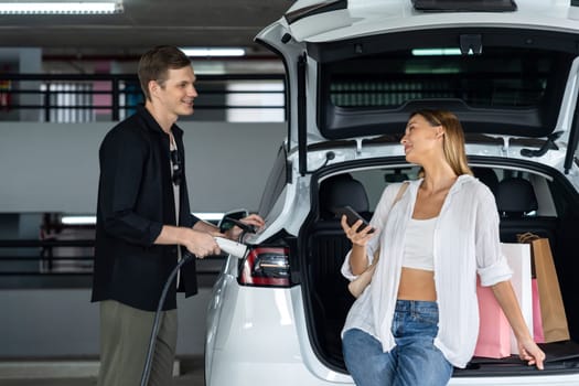 Young couple travel with EV electric car to shopping center parking lot charging in downtown city showing urban sustainability lifestyle by green clean rechargeable energy of electric vehicle innards