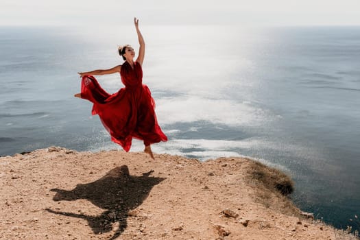 Side view a Young beautiful sensual woman in a red long dress posing on a rock high above the sea during sunrise. Girl on the nature on blue sky background. Fashion photo.