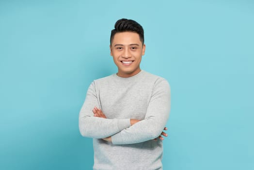 young asian man smiling happy at the camera with arms crossed isolated on blue background