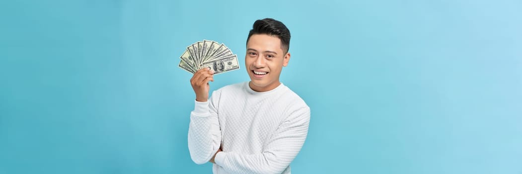 Excited young handsome Asian man holding bunch of money banknotes isolated on blue banner