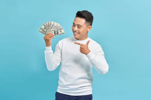 Smiling asian man pointing at fan of money over blue background, copy space