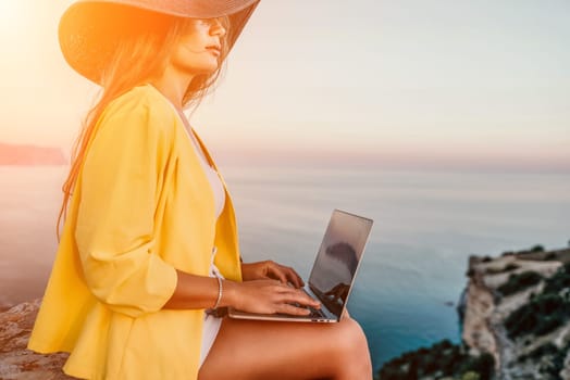 Successful business woman in yellow hat working on laptop by the sea. Pretty lady typing on computer at summer day outdoors. Freelance, travel and holidays concept.