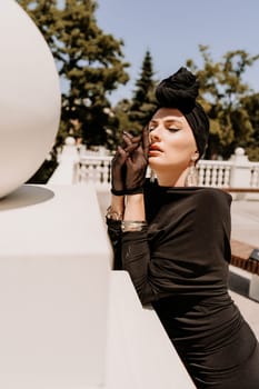 Stylish woman in the city. Fashion photo of a beautiful model in an elegant black dress posing against the backdrop of a building on a city street.