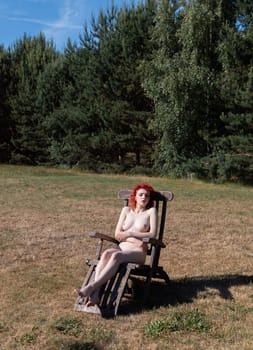 Young woman with scarlet hair poses nude sunbathing in a forest clearing sitting on a wooden country chair