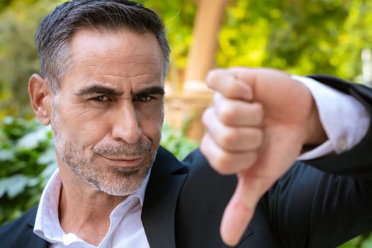 Businessman in a suit outdoors, making a thumbs-down gesture with a focused expression. Captures the concept of disapproval, decision-making, or expressing disagreement in a professional setting.
