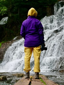 A woman stands with her back to the river in autumn in a yellow suit and purple jacket. The person travels through the Ukrainian Carpathians to the Chervonohorod Dzhuryn waterfall. Vertical