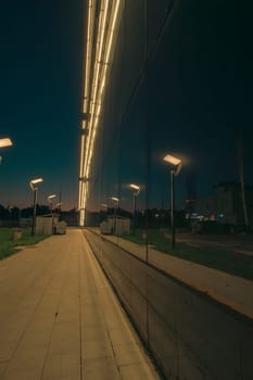 A nighttime view of an illuminated walkway beside a modern glass building with reflective surfaces, creating a sleek and futuristic urban atmosphere