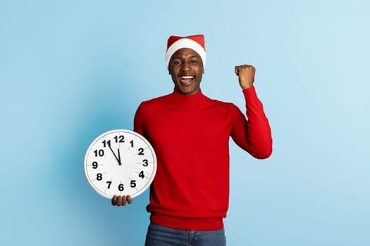 A joyful man dressed in a festive red sweater and a Santa hat holds a clock, showcasing excitement and celebration for the upcoming holiday. The background is a light blue, enhancing the festive mood.