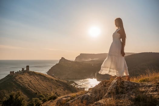 A woman stands on a rocky hill overlooking the ocean. She is wearing a white dress and she is enjoying the view. The scene is serene and peaceful, with the sun shining brightly in the background