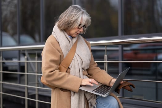 A mature woman, warmly dressed in a coat and scarf, focuses on her laptop while leaning against a railing, surrounded by a contemporary urban environment.