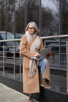 An elderly woman with gray hair is sitting on outdoor steps, focused on her laptop. She wears a cozy overcoat and a scarf, enjoying the autumn breeze while working.