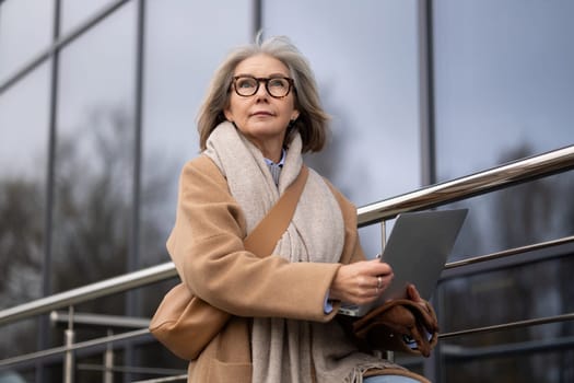A senior woman, dressed warmly in a coat and scarf, sits on a railing outside a contemporary glass building, focused on her laptop as she enjoys her work in the chilly air.