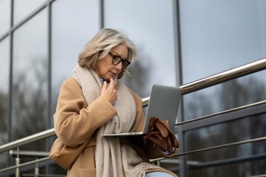 An older woman is focused on her laptop as she sits on a railing outdoors. She is dressed warmly in a coat and scarf, engaged in her work in a contemporary urban setting.