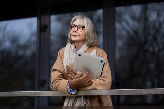 An elderly woman dressed warmly in a coat and gloves stands pensively by a railing. She holds a tablet close, gazing into the distance on a chilly autumn day in the city.
