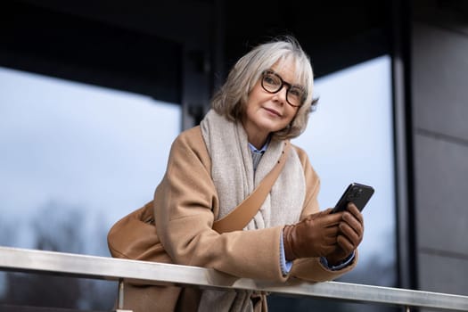 An older woman with gray hair and glasses leans casually against a railing, absorbed in her smartphone. She appears relaxed, dressed warmly for the cool weather.