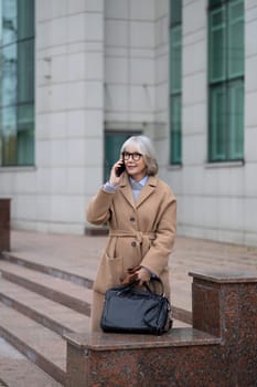 A mature woman with gray hair and glasses is engaged in a conversation on her phone. She stands confidently on stone steps, dressed in a fashionable coat, holding a handbag.