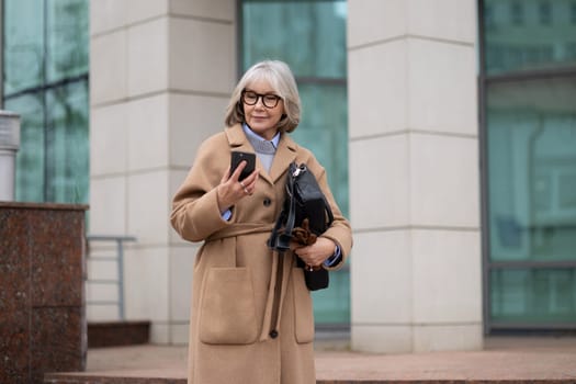 An older woman wearing a beige coat and glasses stands at the entrance of a modern building, looking at her smartphone and carrying a black bag. The weather looks cool.