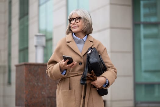 An older woman dressed in a stylish coat and glasses stands confidently outside a contemporary building, looking at her smartphone, enjoying a sunny day.