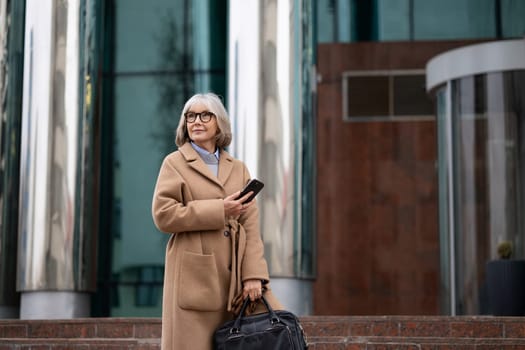A stylish older woman with grey hair stands in front of a contemporary building. She holds a smartphone and a bag, enjoying the outdoors on a bright day.