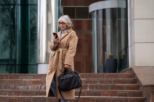 A stylish elderly woman wearing glasses and a beige coat stands on stone steps, holding a smartphone to her ear as she talks. The setting is a contemporary city building on a chilly day.
