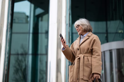 A senior woman with gray hair and glasses stands outside a sleek building while looking at her smartphone. She wears a fashionable beige coat and carries a bag, enjoying the day.