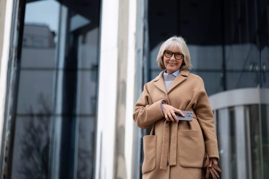 A smiling older woman stands outside a sleek modern building, wearing a fashionable coat and holding a smartphone. The scene captures a sunny day and an urban atmosphere.