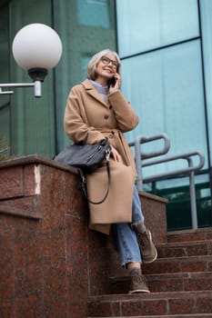 A smiling woman with glasses enjoys a conversation on her phone while seated on outdoor stairs. She carries a bag and is dressed casually in a fashionable coat, showcasing a relaxed vibe.