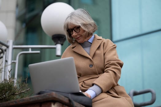 An elderly woman with glasses sits on stone steps, focused on her laptop. She is dressed in a brown coat and enjoys the outdoors while working in a vibrant urban setting.