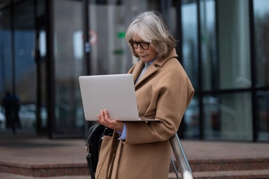 A senior woman with silver hair and glasses stands outside a contemporary glass building, focused on her laptop. She appears engaged in work or communication, enjoying the outdoors.