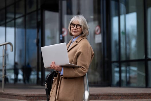 A confident woman with glasses holds a laptop while standing outside a contemporary office building. She is dressed in a stylish, tan coat and appears ready for her workday.