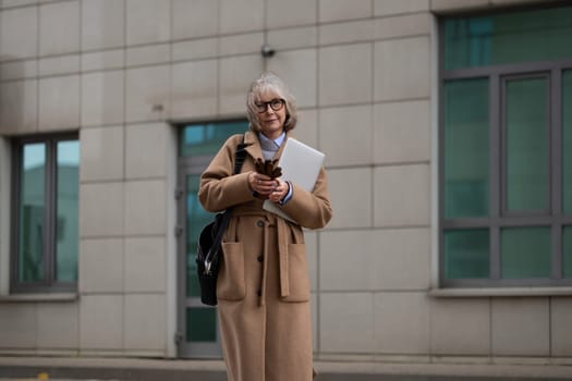 A business woman dressed in a beige coat stands outside a contemporary building while holding a laptop and checking her phone. The day appears overcast, adding a muted atmosphere.