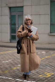An older woman dressed in a fashionable long coat is standing outside a modern building. She holds a laptop and some notes, exuding confidence and style in a chilly environment.