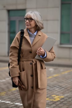 An older woman strolls down the street, wearing a chic coat and carrying a laptop. She exudes confidence and style amidst the modern urban surroundings.