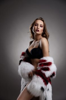 Gorgeous sexy woman wearing a fur coat and luxurious blue velvet lingerie portrait against gray wall