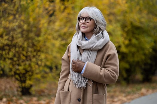 The senior woman stands gracefully in a park filled with colorful autumn leaves, embracing the crisp weather in a stylish coat and warm scarf while enjoying the serene atmosphere.