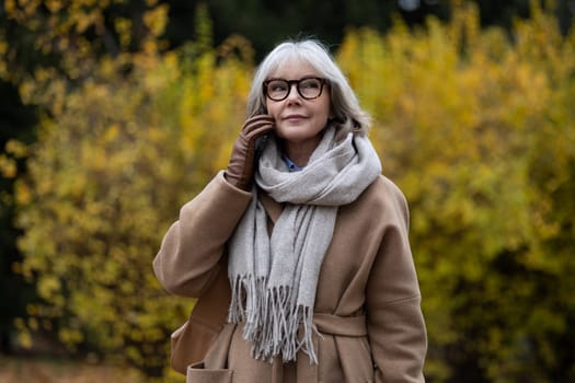 Wearing a warm coat and stylish scarf, a mature woman pauses thoughtfully in a park filled with golden autumn leaves. The serene atmosphere enhances her reflective moment in nature.