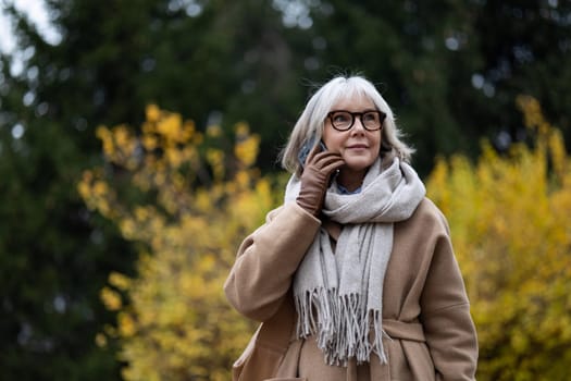 A senior woman stands in a park, wearing a stylish beige coat and a cozy scarf. She smiles while talking on her phone, showcasing the vibrant autumn foliage around her.