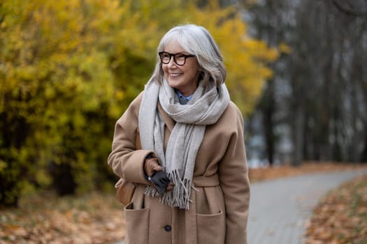 A cheerful elderly woman walks through a park adorned with colorful autumn leaves. Dressed warmly in a coat and scarf, she radiates joy under sunny skies.
