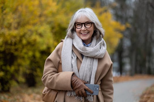 An older woman with gray hair and glasses strolls through a beautiful autumn park. She is smiling, wearing a cozy coat and scarf, holding a smartphone in her hand.