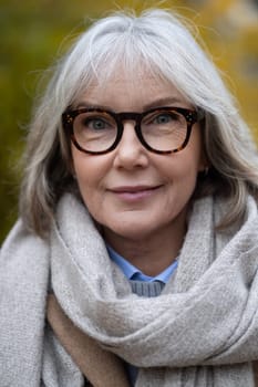 An older woman with gray hair, wearing stylish glasses and a warm scarf, smiles gently while standing outdoors. She enjoys the vibrant colors of autumn in the park.