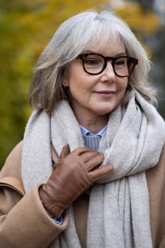 A woman with gray hair wearing glasses and a soft scarf walks through the park on a sunny autumn day, enjoying the vibrant colors of the leaves around her.