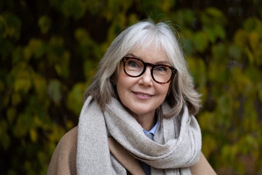 A cheerful older woman stands outside, dressed warmly in a scarf and stylish glasses. The vibrant autumn leaves create a serene backdrop, highlighting her friendly expression.