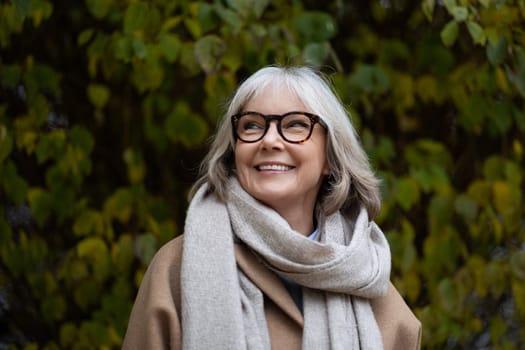 An older woman with gray hair and glasses enjoys the beauty of a sunny autumn day, smiling warmly while surrounded by lush greenery and colorful leaves.
