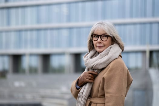 A senior woman poses outdoors in a fashionable coat and scarf, enjoying a sunny day near a contemporary glass building. Her expression reflects confidence and poise.